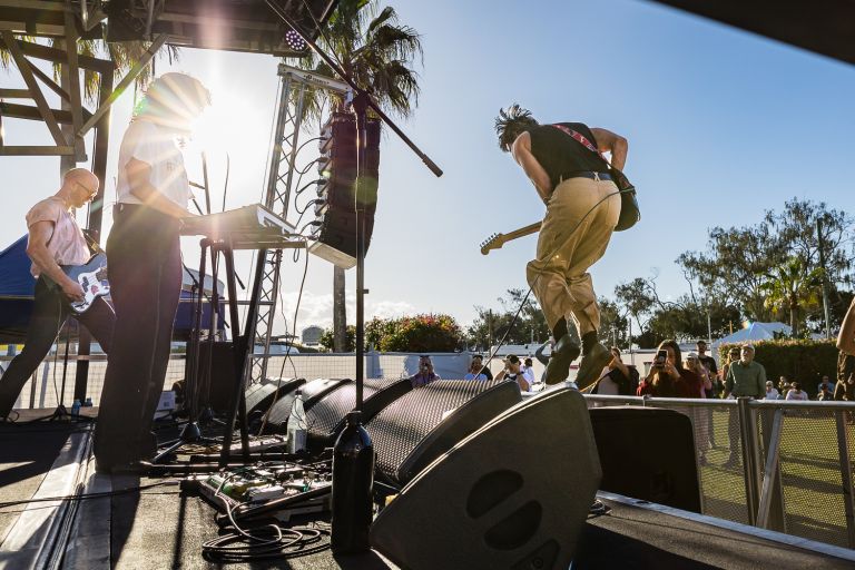 Three musicians on a stage for Byron Music Festival. One musician is jumping in the air with a guitar; another is playing the keyboard, and the third is in the background playing the electric guitar.