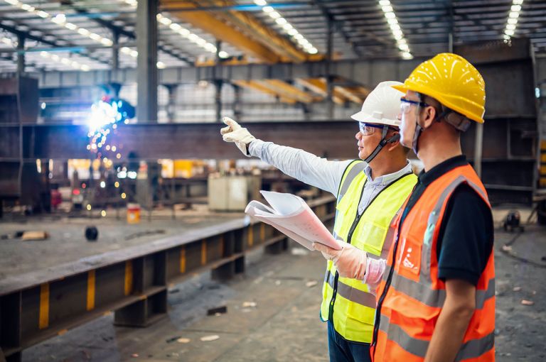 2 workers in hi-vis, holding a document, looking at work being done, inside a production facility