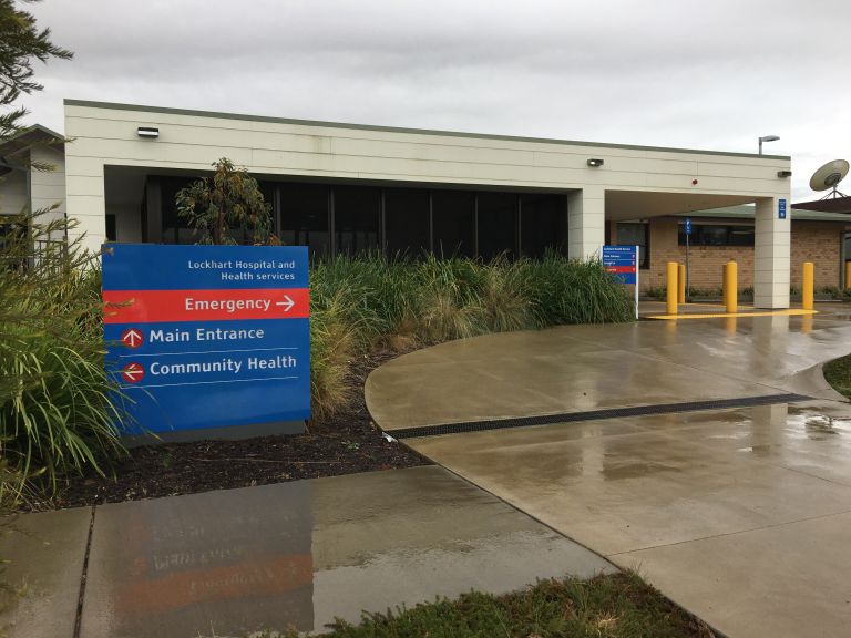 Main entrance to the Lockhart Hospital and Multipurpose Service. A sign to the left of the image shows directions to the main and emergency entry points. A curved concrete driveway curves toward the entrance to the right side of the image.