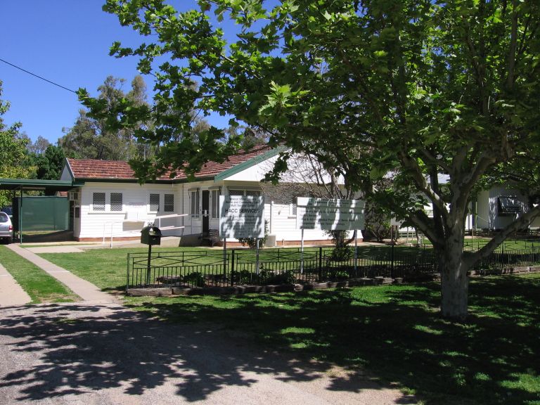 Main entrance to the Moulamein Community Health Centre. The centre's small driveway sits to the left side of the image. A large tree is sited on the verge outside the centre's fenced area. Two white signboards sit on the main lawn of the centre. 