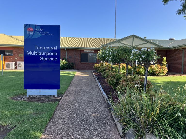 Main entrance to the Tocumwal Multipurpose Service. A walkway leads to the main entrance. To the left of the image, a blue sign with the NSW state logo shows the name of the service and "Murrumbidgee Local Health District". A landscaped green space sits to the right of the walkway. 