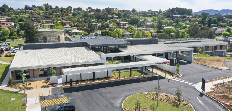 An aerial view of Tumut Health Service. A circular driveway is shown in the bottom-right of the image. Some parking spots, including an accessible parking space sits to the bottom right of the image. The main and emergency entry points is marked on the building. Residences and green spaces spread out behind the building. 