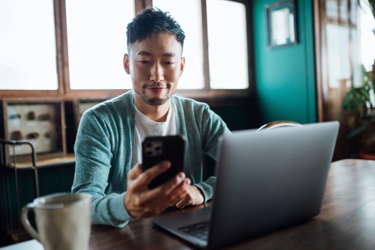 Young man seated looking at phone in hand and laptop open on table.