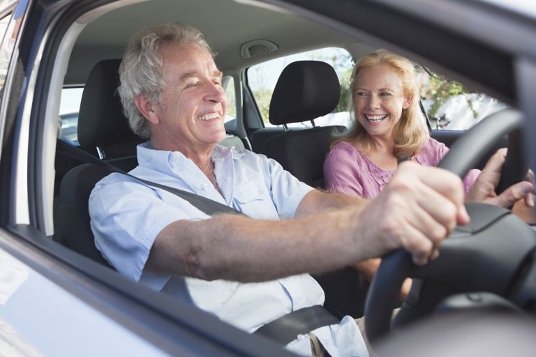 A senior man and woman are sitting in their car. The man is driving.