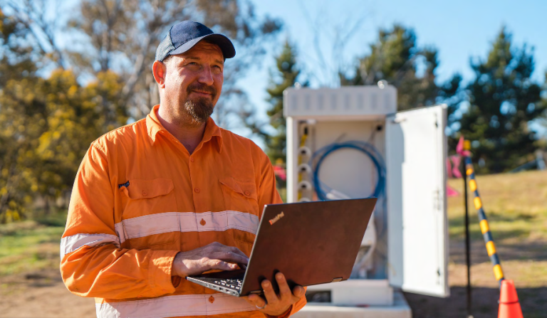 A man wearing a high-visibility shirt and a blue cap holds a laptop in front of a cable box