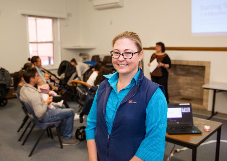 Female allied health worker faces forward and smiles. She is standing in front of a group of new parents at a baby workshop.