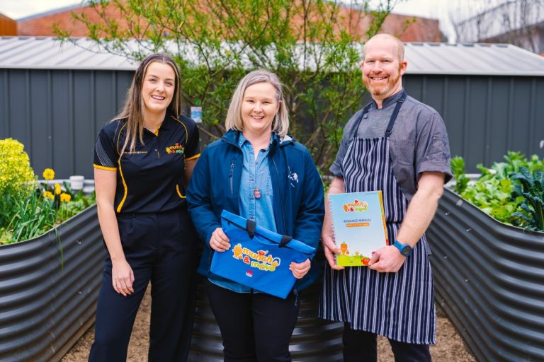 Three healthy eating program staff holding munch-and-move merchandise and standing in front of a community veggie garden