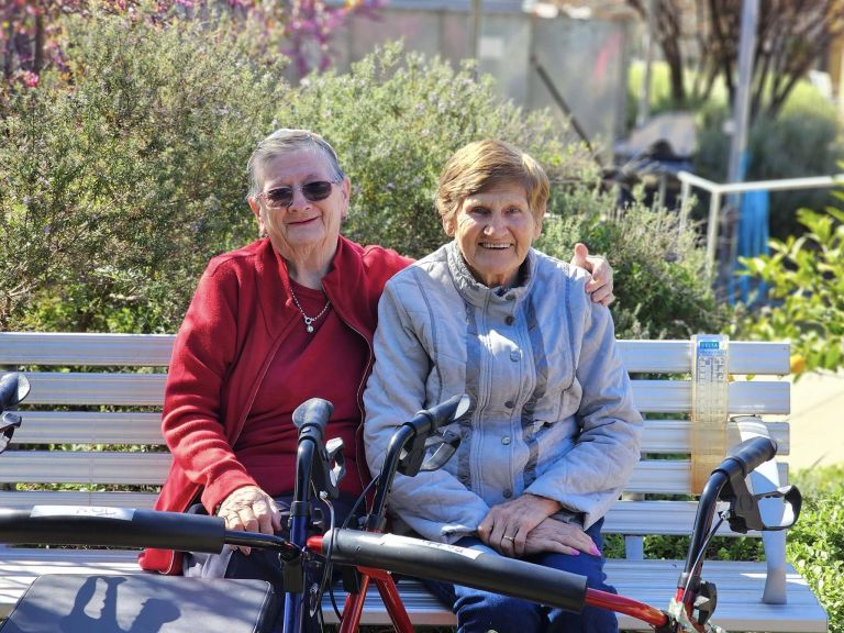 An older woman wearing a red jacket sits with her left arm around another older woman on a park bench. They are facing forward and smiling. A walking aid is in front of them. 