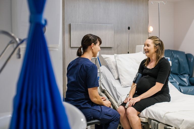 A female nurse sits facing a female patient who is sitting on a hospital bed. They are both facing each other, smiling and talking.