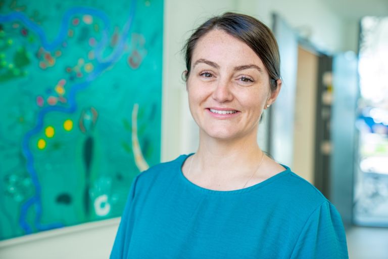 A female healthcare staff faces forward and is smiling. She is standing in a hospital hallway, a painting is in the background toward her left side. 