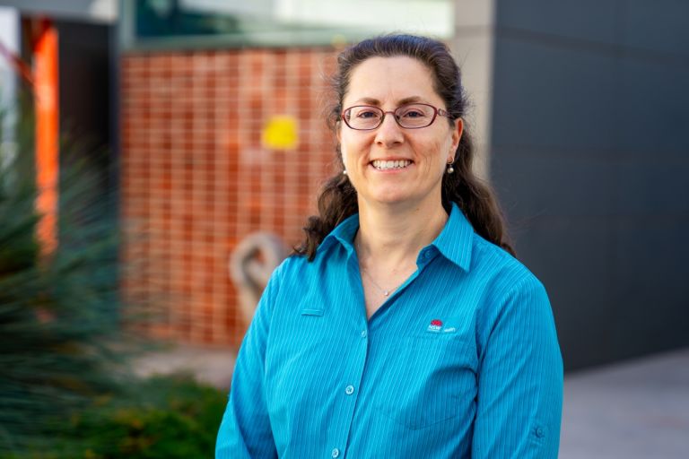 A female allied health worker wearing a blue shirt is standing outside a health facility. She is wearing glasses, facing forward and is smiling.