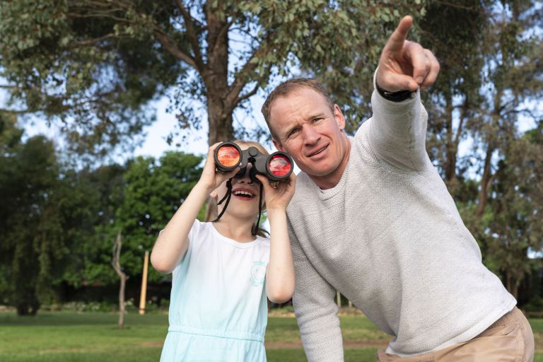 A man wearing a grey long sleeve jumper points to something behind the camera. A young girl in a blue shirt and shorts looks through binoculars she is holding