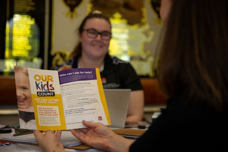 a person holds a flyer with information on the Our Kids Count birth registration program. In the background, a NSW government employee smiles.