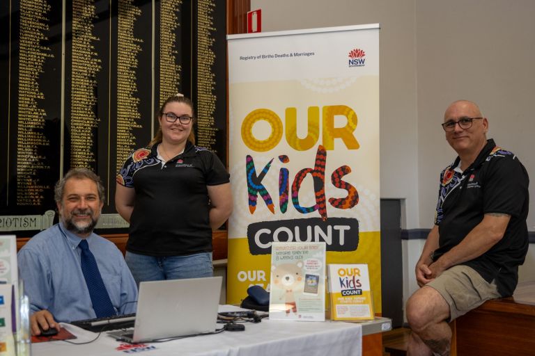 three NSW government staff stand in from of a Poster displaying the text Our Kids Count in bright colours.