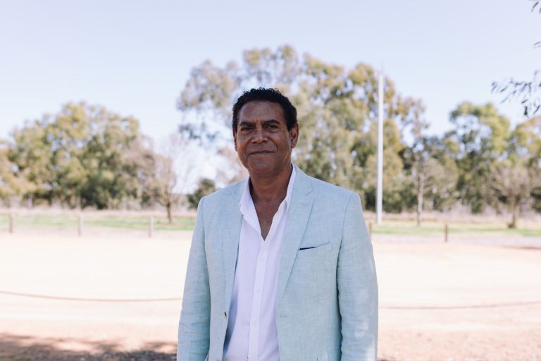 Michael Donovan is relaxed and standing outside with grass, trees and a clear blue sky behind him.