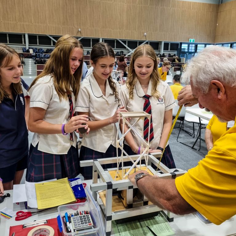 Four teenage girls in school uniform interact with a teacher around a miniature earthquake similator