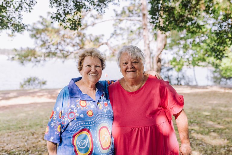 Aunty Gail and Aunty Robyn smile warmly as they stand side-by-side in the shade at a park. Behind them are trees, a lake and clear blue sky.