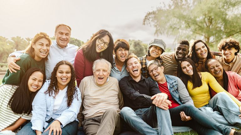 A group of multicultural people sitting close together. They are sitting and kneeling outdoors, facing forward smiling and laughing together.