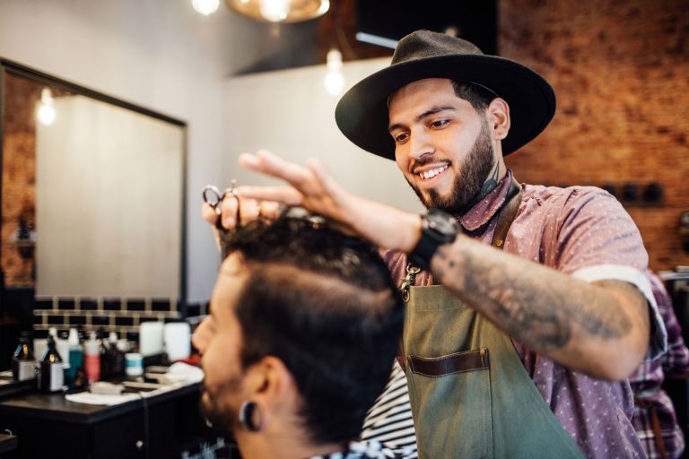 A young hairdresser wearing a hat cuts a customer's hair in a salon.