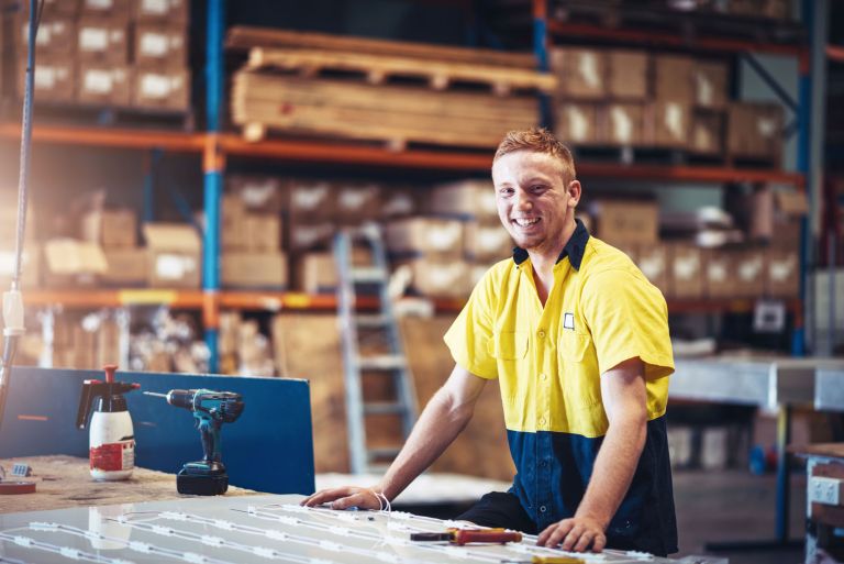 A smiling young man leans on a workbench in a workshop.