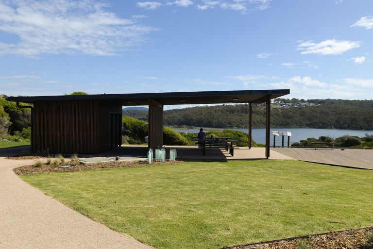 a wooden structure structure with an open awning sites in front of green grass with a path leading out either side. The structure overlooks water and the the sky is blue with some clouds