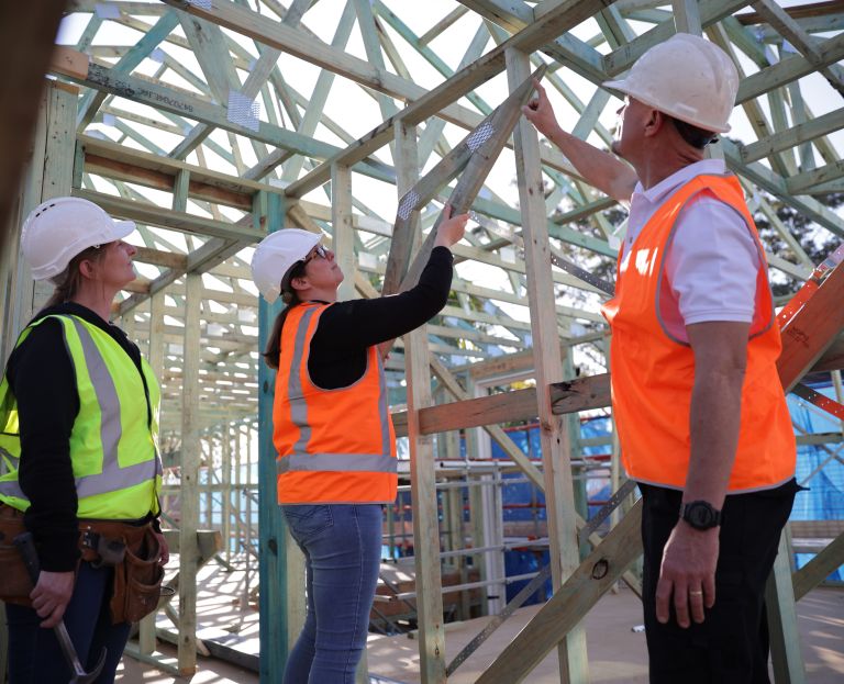 Three people wearing white hard hats and hi-vis vests on site underneath unfinished construction.