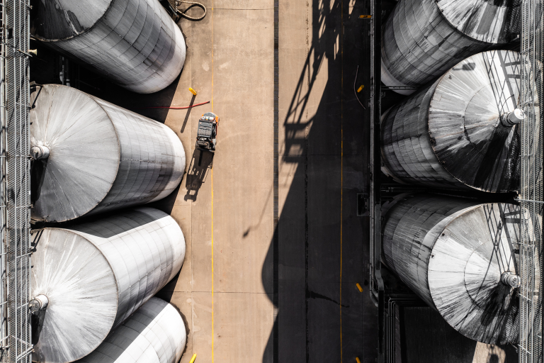 Taken from overhead, the camera looks down on six seven multistorey silver silos. The siloes on the right cast shadows over the concrete they're sitting on. There is a truck parked next to the one of the silos on the left.