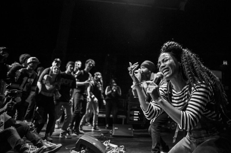 Person wearing striped tshirt and jeans sings into a microphone. There are people in the background. The image is in black and white.