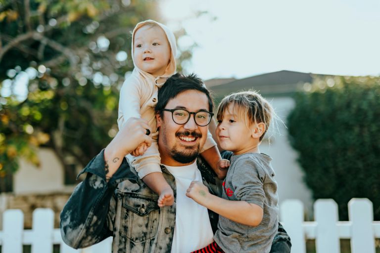 A dad stands outdoors and smiles as he holds his two children.