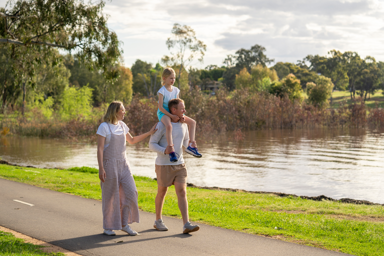 Couple walking on path beside river with girl on their shoulders in Wagga Wagga in the Riverina Murray region