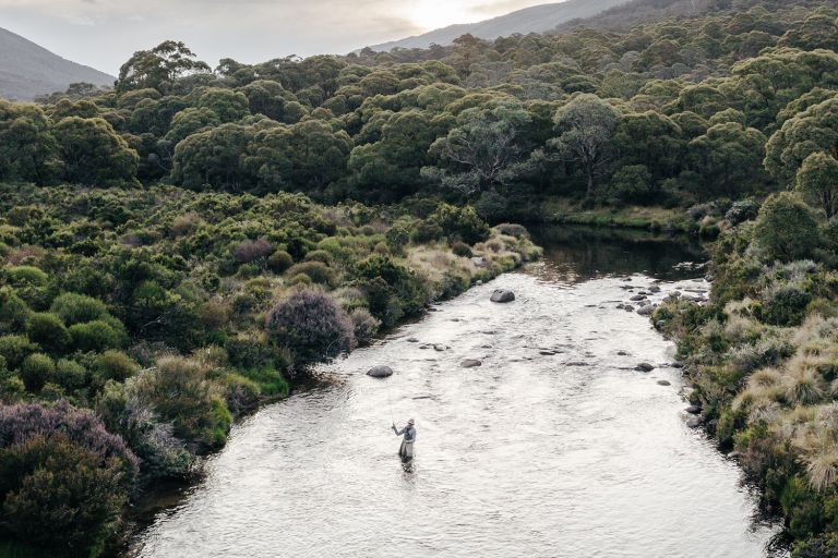 A person stand in the middle of a shallow river fly fishing. The river is surrounded by lush green bush land sloping up to hills. 