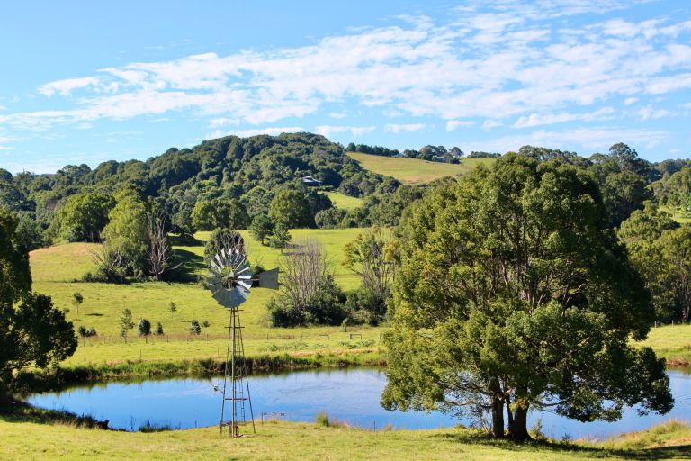 Horizontal landscape in rural country Bangalow with rolling green hills windmill lake and large trees against pastel blue cloudy sky Australia.