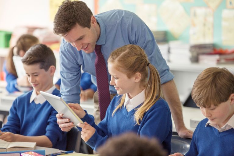 A female student in a classroom with her peers sitting next to her and her male teacher helping her on an iPad