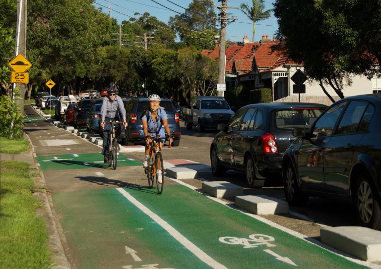 Two people riding bikes on a cycle way beside a road and parked cars