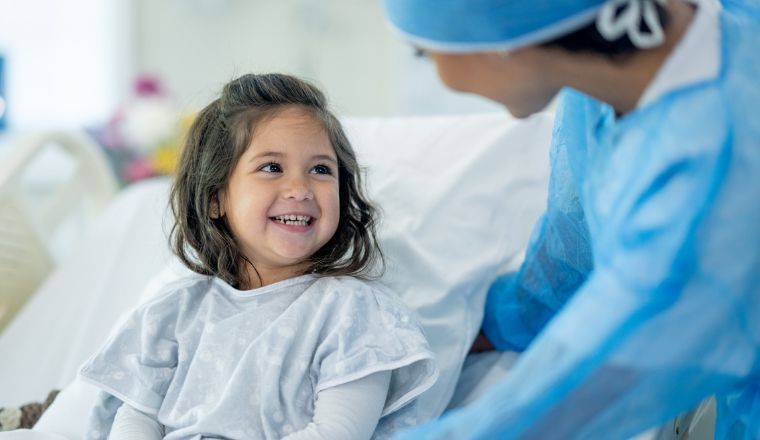 A young girl sitting on her hospital bed speaking to her doctor wearing a surgical gown and cap