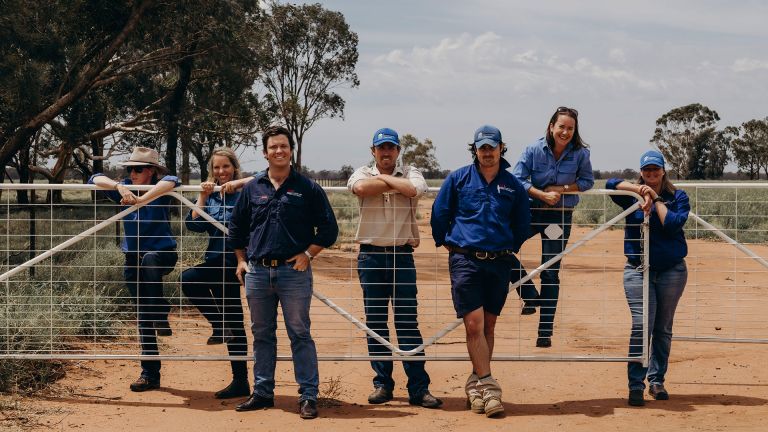 7 lls workers by the gate of a farm on a dirt road smiling for the camera