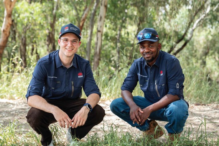 2 lls graduates squatting down and smiling at the camera against a sunny bushland background