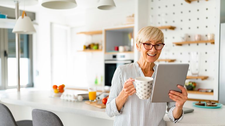 Smiling mature woman drinking coffee while reading online news on digital tablet and leaning on table in the morning.