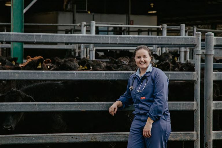 woman smiling at the camera leaning on a metal fence with cattle behind her