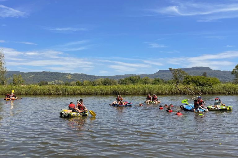 A group of about 20 young people are paddling on circular floatation devices on a lake. Some people are in the water and others are in pairs on the devices. All are wearing life jackets. in the background is some tall grass, trees and then a small ridge of mountains. The sky is bright blue