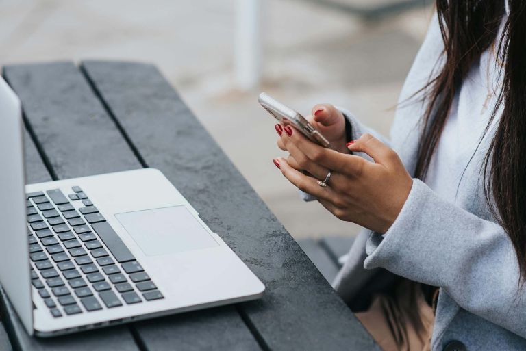 Female holding a mobile phone while sitting at a table with a laptop. 