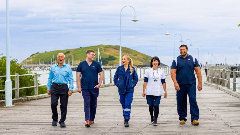 Five people walk along a wooden pier with street lights and a river in the background. The people are in uniforms, showing they are essential workers including doctors and nurses.