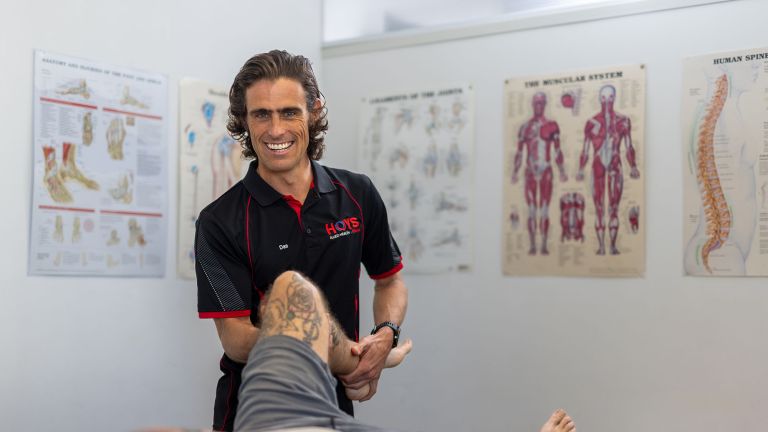 A man smiles at the camera. He is holding someone's leg, who is laying on a physiotherapy table in front of him. There are posters of ligaments, bones and skeletons on the walls behind him