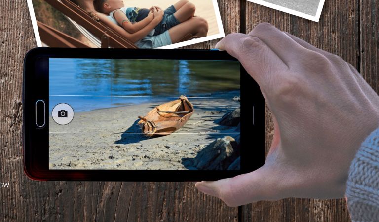 A hand holding a smartphone, capturing a photo of a small dog lying on a sandy beach, with other printed photos scattered on the wooden surface beneath the phone.