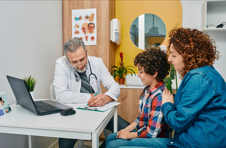 Photo of an older male doctor in consultation rooms with a young boy and his mother, he is writing on a form.