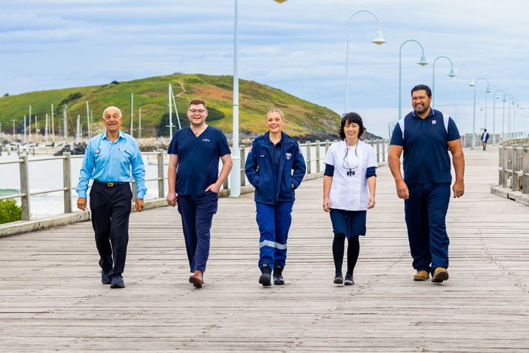 5 essential workers walking across a timber bridge in Coffs Harbour with small boats, a hill and blue sky with streaky clouds in the background