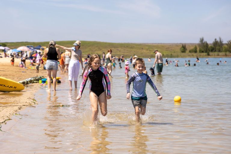 Young boy and girl running thru large body of water at Penrith Beach with families in the background enjoying the beach.