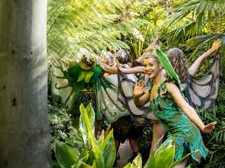 Performers dressed as fairies in leafy costumes and wings walk through a lush, tropical garden.