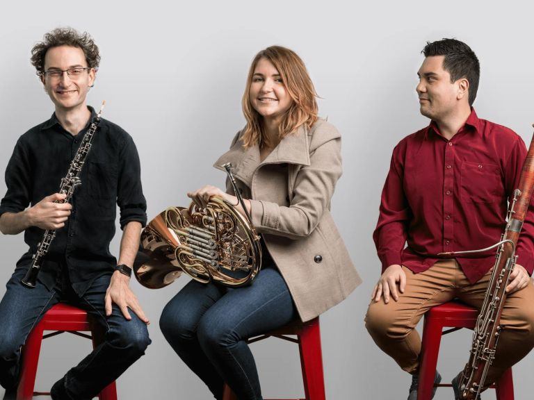 A colour photograph of the members of Arcadia Winds sitting on stools and holding their instruments.