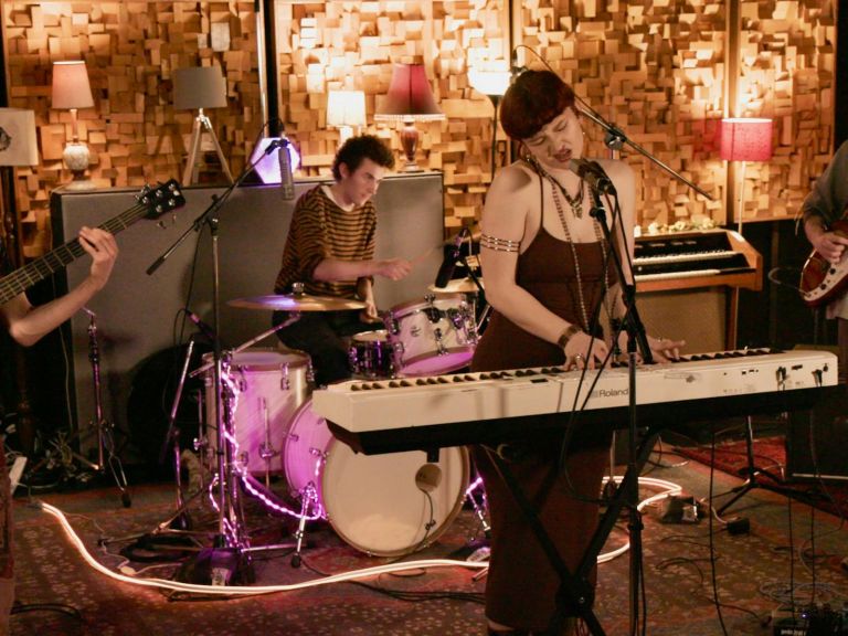 Members of a band standing on a small stage, the singer is a woman in front of a keyboard